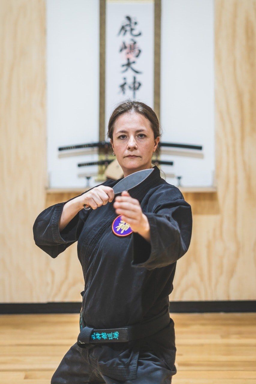 Viviana en keikogi negro dentro del dōjō, frente al kamidana, blandiendo un kunai